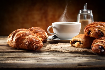 Na dřevěném stole leží croissanty a rohlíky, vedle šálek kávy a mlýnek s párou.